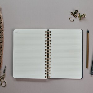 May include: An open notebook with lined pages, a gold spiral binding, and the word "NOTES" at the top. A feather, scissors, a pencil, a ruler, paper clips, and a tassel are arranged around the notebook on a gray surface.