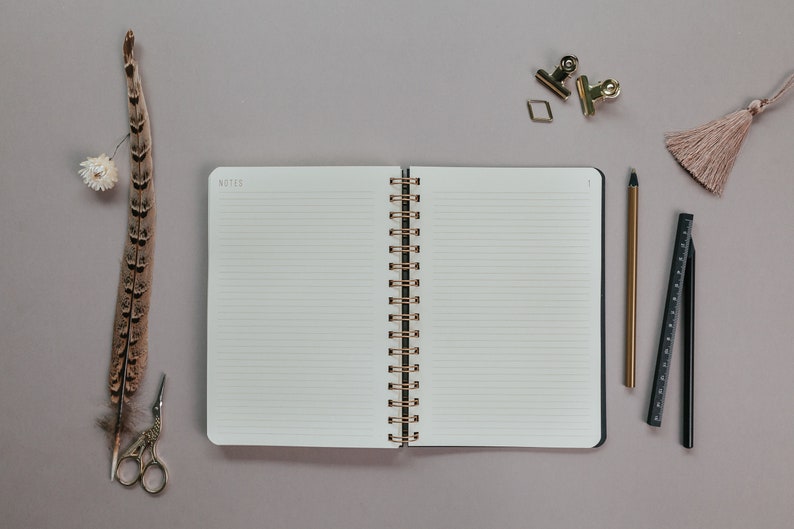 May include: An open notebook with lined pages, a gold spiral binding, and the word "NOTES" at the top. A feather, scissors, a pencil, a ruler, paper clips, and a tassel are arranged around the notebook on a gray surface.