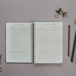 May include: An open spiral-bound planner for the year 2021, displaying monthly calendars. The planner is surrounded by stationery items, including a gold pen, a ruler, and paper clips. A feather and tassel add decorative elements.