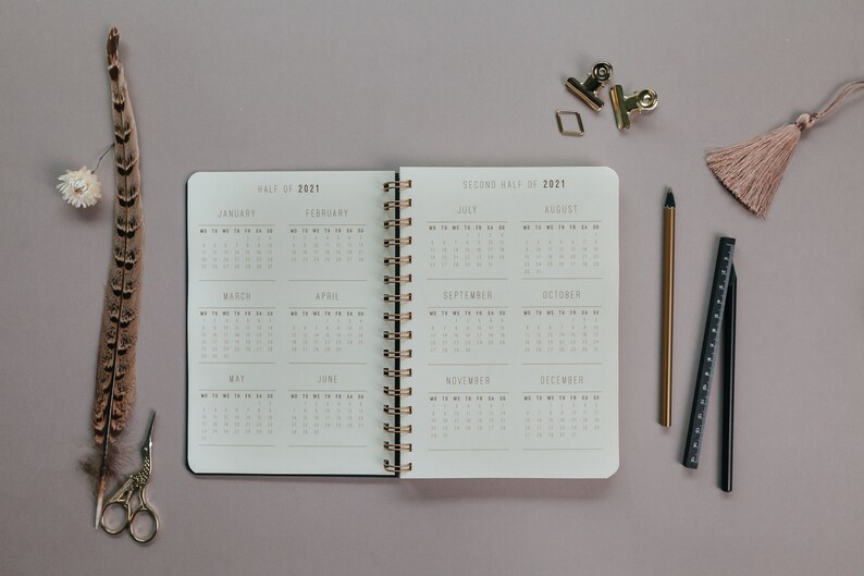 May include: An open spiral-bound planner for the year 2021, displaying monthly calendars. The planner is surrounded by stationery items, including a gold pen, a ruler, and paper clips. A feather and tassel add decorative elements.
