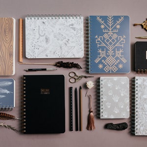 May include: An assortment of spiral-bound notebooks and planners with various cover designs, including wood grain, botanical patterns, and geometric shapes. Accessories like pens, scissors, and decorative elements are also present.