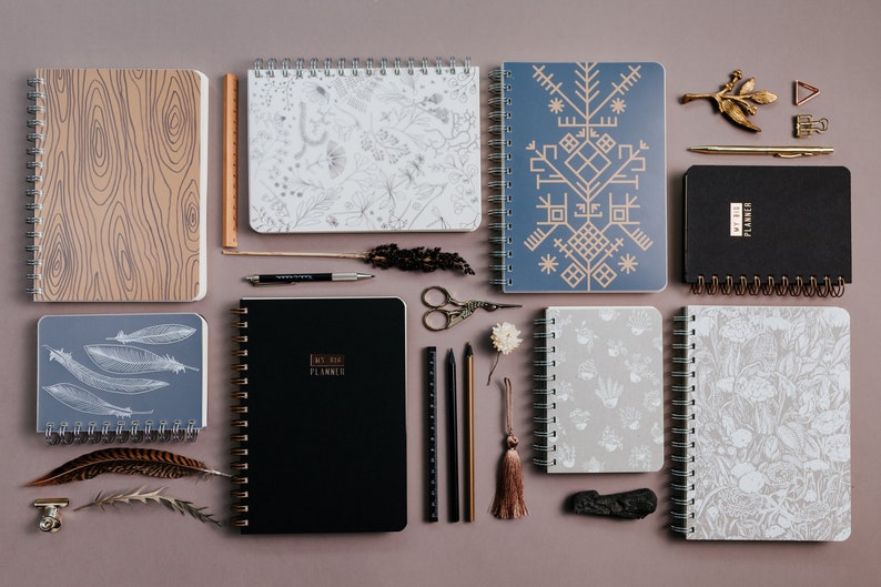May include: An assortment of spiral-bound notebooks and planners with various cover designs, including wood grain, botanical patterns, and geometric shapes. Accessories like pens, scissors, and decorative elements are also present.