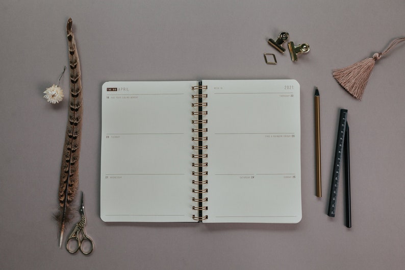May include: Open weekly planner with gold spiral binding, featuring the month of April 2021. Includes a gold pen, black ruler, paper clips, and a decorative tassel. A feather and small scissors are also present.
