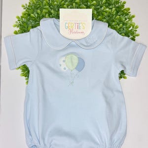 Peut inclure: Une barboteuse à manches courtes bleu clair avec un col claudine à passepoil blanc et un motif de ballons brodés aux motifs pois, uni et vichy. Le vêtement comporte une étiquette portant l'inscription Gertie's Heirlooms.