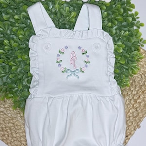 Pode incluir: Macacão branco para bebé com detalhes de babados e grinalda floral bordada com laço rosa e a letra "A". O macacão tem alças ajustáveis e fechos de botão. O texto "Gertie's Heirlooms" está no topo.