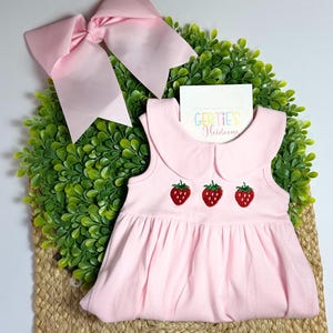 May include: A light pink baby dress with a Peter Pan collar and three embroidered strawberries. A large pink bow is positioned above the dress. The dress is displayed on a green wreath.