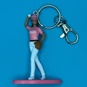 May include: A Barbie keychain featuring a baseball player figurine in a pink uniform with the word "Barbie" and the number "59". The figurine is holding a baseball and a glove, with a silver keyring and clasp.