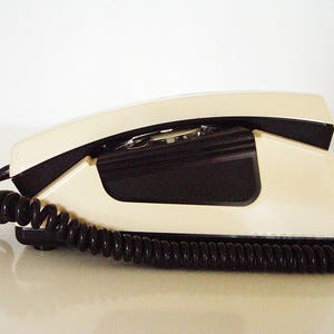 Puede incluir: Un tel&eacute;fono de disco vintage de dise&ntilde;o blanco y negro. El tel&eacute;fono tiene un cable en espiral y un auricular.