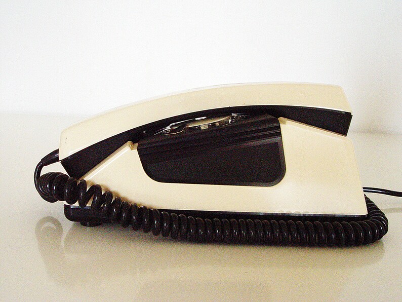 Puede incluir: Un tel&eacute;fono de disco vintage de dise&ntilde;o blanco y negro. El tel&eacute;fono tiene un cable en espiral y un auricular.