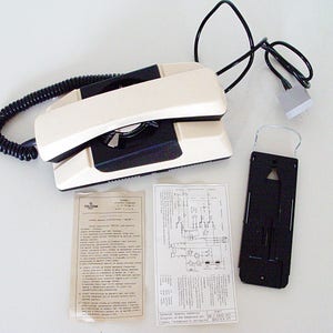Puede incluir: Un tel&eacute;fono rotatorio blanco y negro con un cable en espiral y un soporte de pared. El tel&eacute;fono tiene un dial negro y un auricular blanco. El soporte de pared es negro y tiene un gancho de metal.