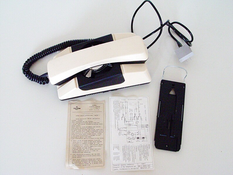 Puede incluir: Un tel&eacute;fono rotatorio blanco y negro con un cable en espiral y un soporte de pared. El tel&eacute;fono tiene un dial negro y un auricular blanco. El soporte de pared es negro y tiene un gancho de metal.