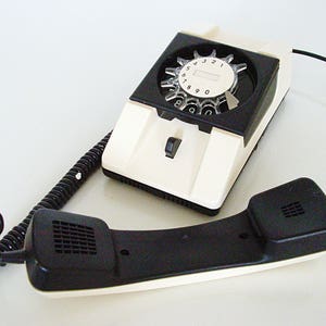 Puede incluir: Un tel&eacute;fono de disco blanco con un disco negro y un auricular negro. El tel&eacute;fono tiene un cable blanco.