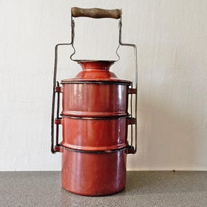 May include: A vintage red metal lunch carrier with three stacked compartments. It has a metal handle with a wooden grip. The carrier is designed to hold food and is likely from an earlier era.