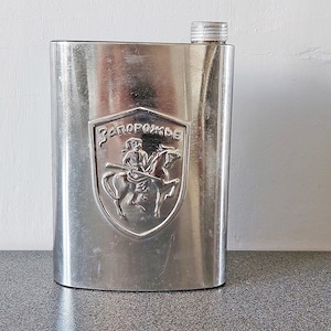 May include: A silver metal flask with a screw-on cap. The flask has a raised, embossed shield with the text "Запорожье" and a depiction of a knight on horseback.