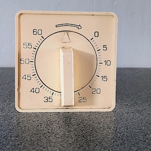 Pode incluir: Um temporizador de cozinha vintage creme, com formato quadrado. Possui um mostrador preto com números de 0 a 60, um botão central e uma seta preta. O temporizador está sobre uma superfície escura. Medidas em minutos.