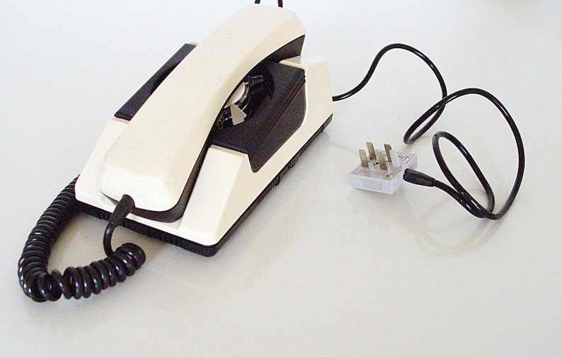 Puede incluir: Un tel&eacute;fono rotatorio blanco con un dial negro y un cable en espiral. El tel&eacute;fono tiene un auricular blanco y una base negra. El tel&eacute;fono est&aacute; conectado a un adaptador de corriente blanco con un cable negro.
