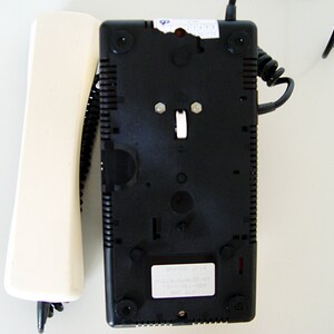 Puede incluir: Un tel&eacute;fono rotatorio negro con un auricular blanco. El tel&eacute;fono tiene un cable negro y un bot&oacute;n blanco en la parte delantera. El tel&eacute;fono est&aacute; sobre una superficie blanca.