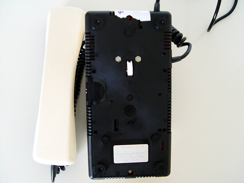 Puede incluir: Un tel&eacute;fono rotatorio negro con un auricular blanco. El tel&eacute;fono tiene un cable negro y un bot&oacute;n blanco en la parte delantera. El tel&eacute;fono est&aacute; sobre una superficie blanca.