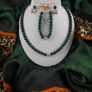 May include: A green malachite necklace, bracelet, and earrings set with gold accents. The jewelry is displayed on a white stand against a green and brown patterned fabric.