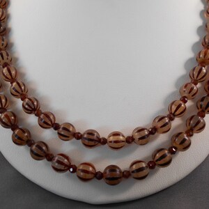 Puede incluir: Un collar de dos hilos con cuentas a rayas marrones y beige. Las cuentas redondas tienen un diseño acanalado. El collar se exhibe sobre un busto blanco, mostrando el diseño y el color de la joya.
