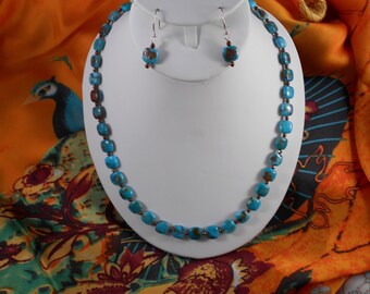 May include: A turquoise bead necklace and matching earrings set displayed on a white bust. The necklace and earrings feature square turquoise beads with brown accents. The jewelry is set against a colorful patterned fabric background.