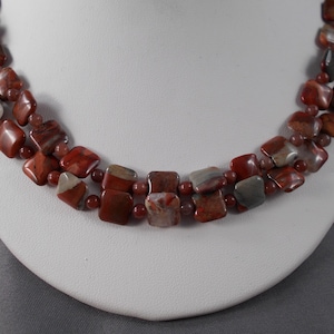 Puede incluir: Un collar de doble cadena hecho de cuentas de jaspe rojo, marrón y blanco. Las cuentas son de forma cuadrada y tienen un acabado pulido.