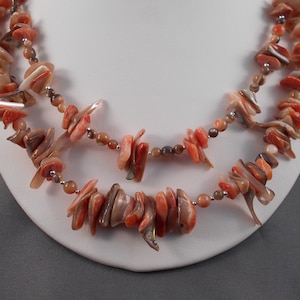 Puede incluir: Un collar de doble cadena hecho de cuentas de concha natural en tonos rosa, naranja y marrón. Las cuentas son irregulares en forma y tamaño, lo que le da al collar un aspecto único y orgánico.