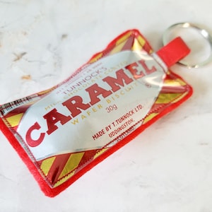 May include: A rectangular keychain featuring a Tunnock's Caramel Wafer Biscuit wrapper design in red, gold, and silver. The text reads Tunnock's Caramel Wafer Biscuit, 30g, Made by T. Tunnock Ltd. Uddingston, Scotland. It has a red felt backing and a metal ring.