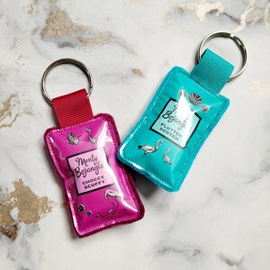 May include: Two keychains with rectangular pouches. One is magenta with the text "Monty Bojangle Choccy Scoffy" and a flamingo illustration. The other is teal with "Bojangle Flutter Scotch" and swan illustrations. Both have silver keyrings.