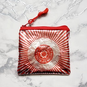 May include: A red and silver zippered pouch with a red pull tab. The pouch is decorated with a white and red striped pattern. The pouch has a white label with the text "Tunnack's Milk Chocolate Mallow Biscuit 249g" printed on it.