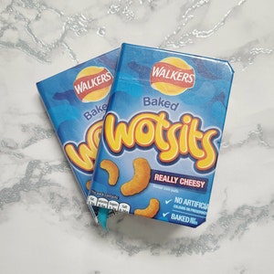 Può includere: Due confezioni blu e gialle di snack Walkers Baked Wotsits. Le confezioni presentano il logo del marchio e le parole "Baked Wotsits REALLY CHEESY". Sono su una superficie di marmo bianco e grigio.