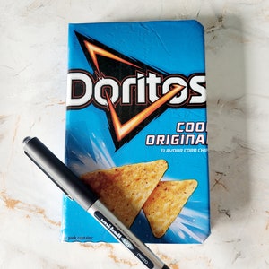 Op de afbeelding: Een blauwe Doritos Cool Original maïschipszak met een zwarte en zilveren Uni-ball Eye Micro pen. De zak toont het Doritos-logo en afbeeldingen van de chips. De pen ligt diagonaal over de zak.