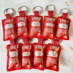 May include: Eight red felt keychains shaped like Taco Bell hot sauce packets. Each keychain has a different funny saying on the front, such as "Too bad you're not single" and "Marry me!"