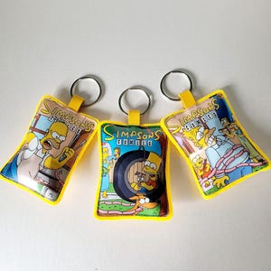 Może przedstawiać: Trzy prostokątne breloki z grafikami Simpsons Comics, żółtymi obwódkami z tkaniny i metalowymi kółkami. Każdy brelok przedstawia kolorowe sceny z kreskówek z postaciami takimi jak Homer, Bart i Marge oraz logo Simpsons Comics.