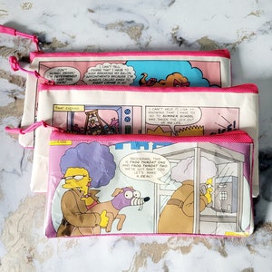 May include: Three canvas pencil cases with pink zips and comic strip designs. The cases feature panels with text and cartoon characters. The top case reads "Don't worry, Pierre! I'm determined to keep this date!"