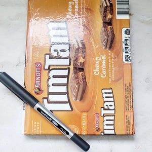 Può includere: Una scatola di biscotti al cioccolato e caramello morbido Tim Tam di Arnott, con una penna nera. La scatola è arancione e marrone, con le parole "Tim Tam" e "Chewy Caramel" visibili. Peso netto 175 g.