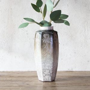 May include: A white ceramic vase with a crackled glaze and a geometric design. The vase is filled with green leaves.