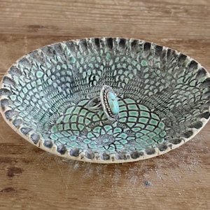 May include: Oval ceramic dish with a textured, patterned surface in shades of teal, gray, and black. A silver ring with a turquoise stone rests inside the dish. The dish has a decorative, scalloped edge.