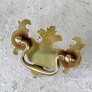 May include: A gold-toned ornate drawer pull with a decorative, scrolled design.