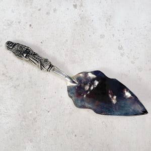 May include: A silver cake server with an ornate handle featuring a detailed figure. The blade has a curved, leaf-like shape. The server is displayed on a light gray, speckled surface.