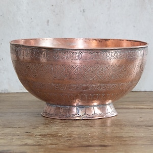 May include: A copper bowl with an embossed floral pattern. The bowl has a wide, rounded shape and sits on a small, decorative base. The copper has a warm, reddish-brown hue, and the embossed details add texture.