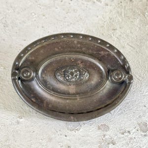 May include: An oval-shaped, silver-toned metal drawer pull with a decorative design. The pull has a raised, oval-shaped center with a floral design.