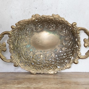 May include: An antique brass oval tray with ornate openwork detailing and two curved handles. The tray has a tarnished, aged appearance with hints of green oxidation, suggesting its vintage. The decorative design features floral and scroll patterns.