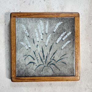 May include: Square decorative tile with a wooden frame. The tile features a stylized illustration of wheat stalks in white and blue against a gray background. The wooden frame is a warm brown color.