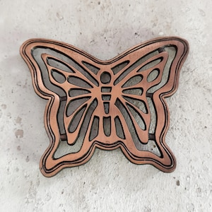 May include: A copper-colored butterfly-shaped decorative object. The butterfly design features an intricate openwork pattern with a dark brown outline. The object is likely made of metal and is displayed against a neutral, textured background.