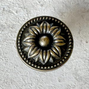 May include: Antique brass-toned cabinet knob with a sunflower design. The knob features a detailed floral center surrounded by a beaded edge. This decorative hardware adds a vintage touch to furniture and cabinets.