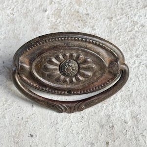 May include: Antique bronze oval drawer pull with a floral center design and a curved handle. The hardware is aged with visible wear and tear, showcasing its vintage character. The pull measures approximately 4 inches in length.