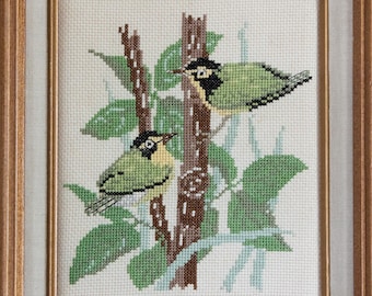 May include: Cross-stitch artwork of two birds perched on a tree branch with green leaves. The birds have yellow and black markings, and the tree trunk is brown. The artwork is framed in a wooden frame.