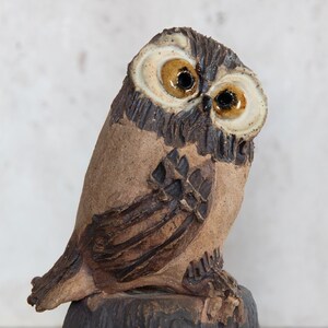 May include: A ceramic owl figurine with a tilted head. The owl has brown and tan coloring with detailed feather patterns. The eyes are a striking combination of brown and yellow. The owl is perched on a dark, textured base.