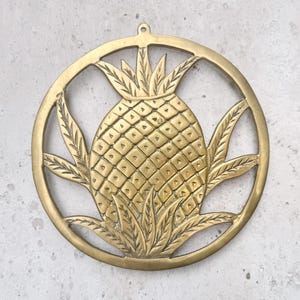 May include: A gold-toned metal pineapple wall decor with a circular shape and intricate details. The pineapple is surrounded by leaves and has a textured surface.